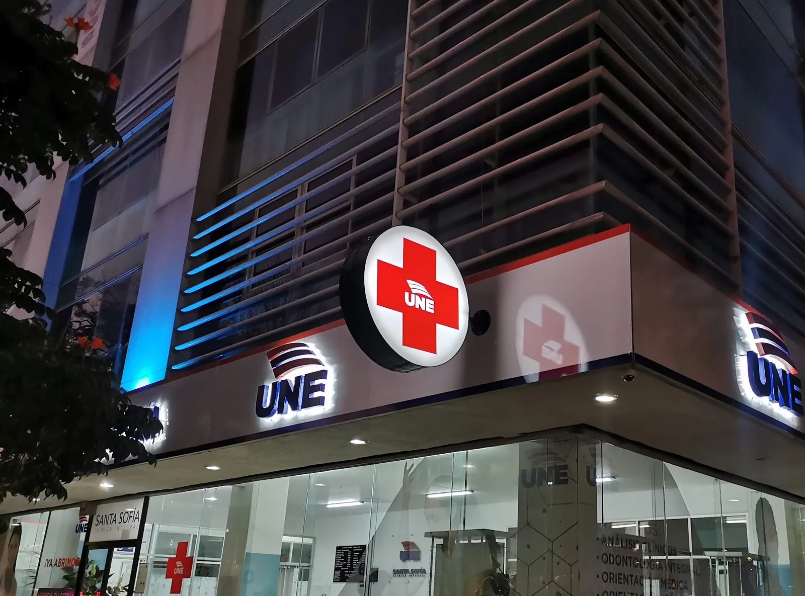 Fachada de una clínica UNE con cruz y letras iluminadas, al anochecer